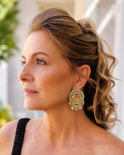 Woman wearing colorful earrings with a blurred background-Pearl-gold-handmade-crochet-drop-earrings-Minerva-Duo-style- 18k-gold-filled-wire-lightweight-hypoallergenic- Handmade in Brazil, these double link hand crocheted mesh earrings feature 18 karat gold filled wire, pearly crystals, lightweight comfort, and hypoallergenic materials.