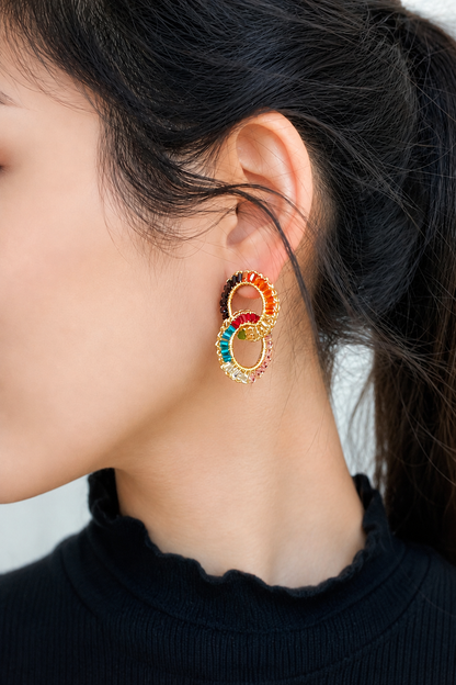Close-up of a person wearing colorful hoop earrings with a blurred background. Handmade in Brazil, these small double link hand crocheted earrings feature 18 karat gold filled wire, multicolor crystals, lightweight comfort, and hypoallergenic nickel free materials.