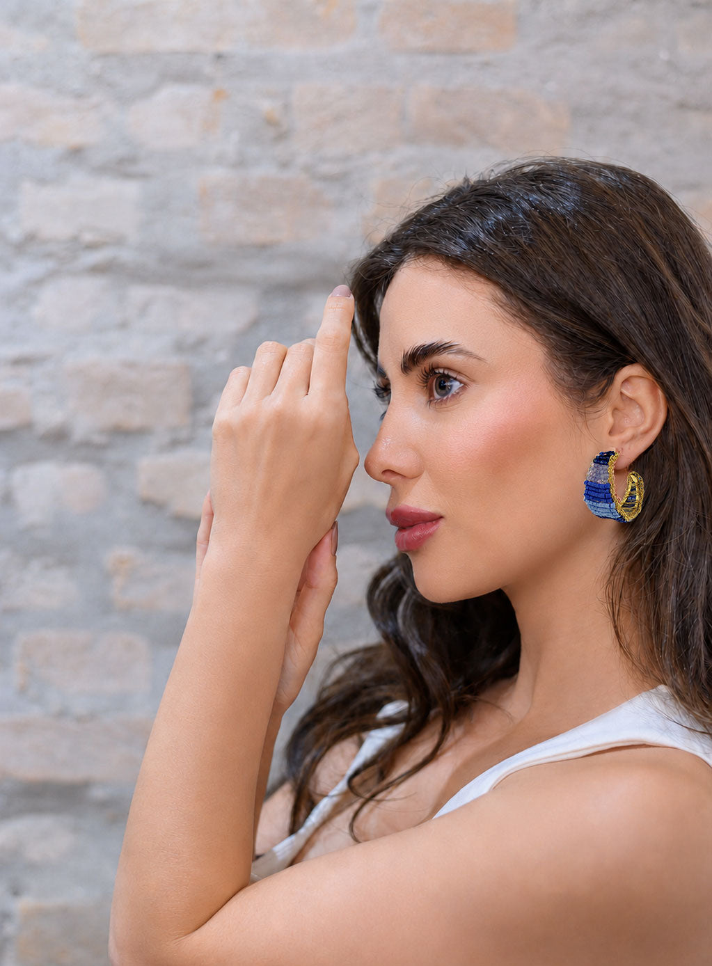 Woman wearing blue earrings against a stone wall background. Vibrant handmade crochet hoop earrings with enameled wire and crystals, handcrafted in Brazil by skilled artisans.