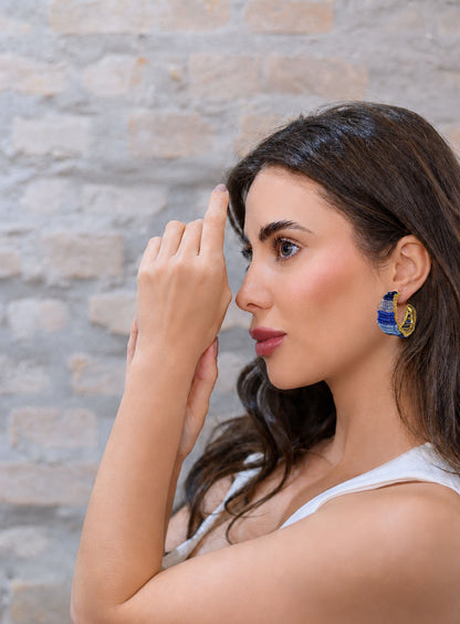 Woman wearing blue earrings against a stone wall background. Vibrant handmade crochet hoop earrings with enameled wire and crystals, handcrafted in Brazil by skilled artisans.