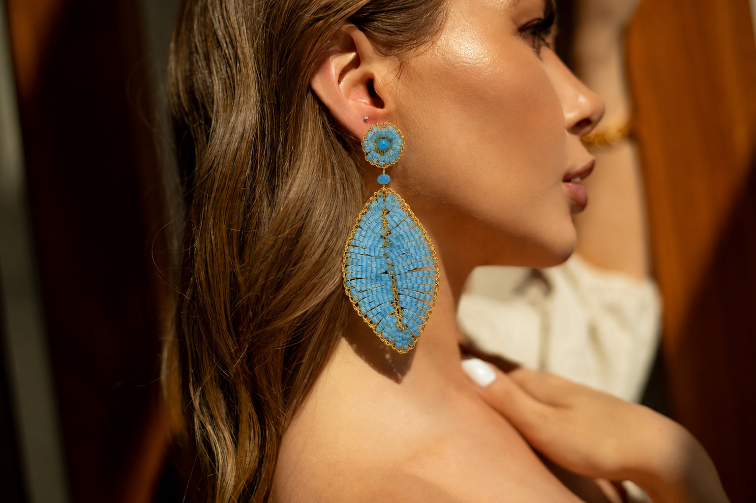 Close-up of a woman wearing blue beaded earrings with a blurred background. Leaf-shaped hand-crocheted dangle earrings in 18 karat gold filled wire with aqua blue crystals and beads, handmade in Brazil, lightweight hypoallergenic nickel-free artisan jewelry.