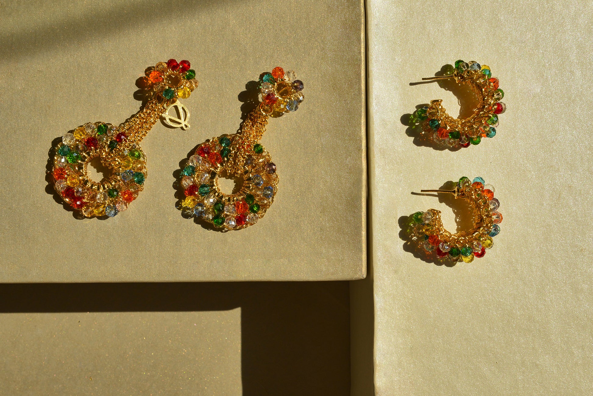 Gold hoop earrings with colorful beads on a beige background. Handmade in Brazil, these hand crocheted earrings feature 18 karat gold filled wire, multicolor crystals, lightweight comfort, and hypoallergenic nickel free materials.
