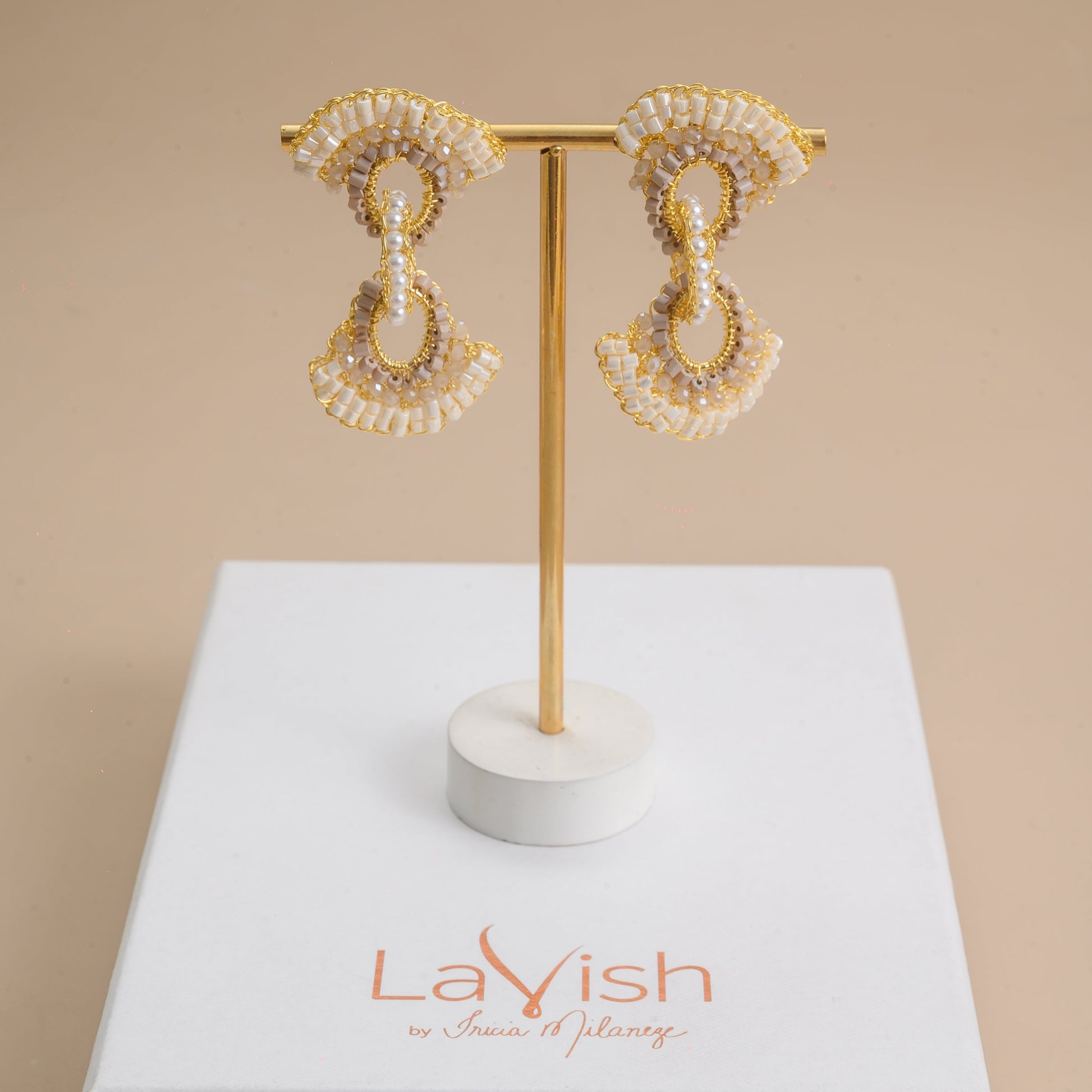 Pair of gold and pearl earrings on a stand with 'LaVish' branding on a beige background