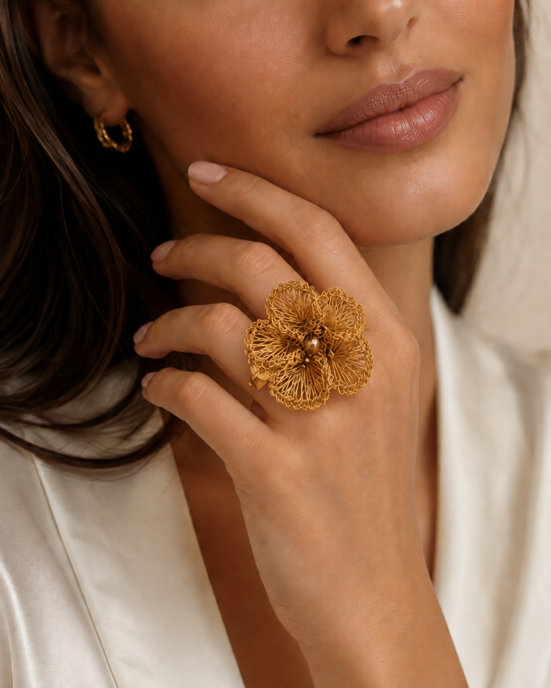 Close-up of a woman's hand wearing a gold floral ring with a neutral background. Floral hand-crocheted adjustable ring in 18 karat gold filled wire, handmade in Brazil, hypoallergenic nickel-free artisan statement jewelry.