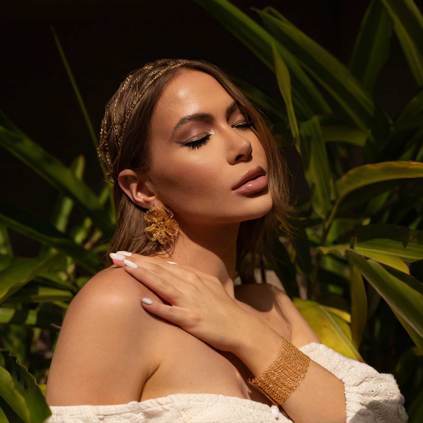 Woman in a white off-shoulder top standing among green foliage - Handmade in Brazil, these floral hand crocheted earrings feature 18 karat gold filled wire, lightweight comfort, and hypoallergenic nickel free materials. 8202-001-000 504-0001-000