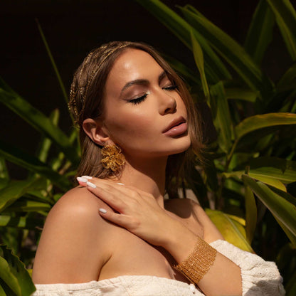 Woman in a white off-shoulder top standing among green foliage - Handmade in Brazil, these floral hand crocheted earrings feature 18 karat gold filled wire, lightweight comfort, and hypoallergenic nickel free materials. 8202-001-000 504-0001-000