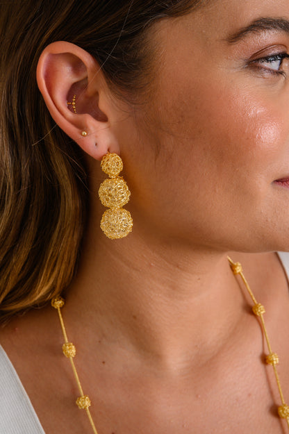 Close-up of a woman wearing gold earrings with a blurred background. Hand-crocheted 18k gold filled earrings handmade in Brazil, lightweight hypoallergenic nickel-free artisan wire crochet earrings. 
