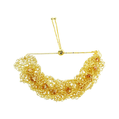 A gold-toned statement collar necklace featuring a dense, textured mesh of twisted chain links with small faceted amber-orange crystal accents, suspended from a delicate adjustable chain with a central gold bead. Shop the Ambar & Gold Calla Embrace Bracelet. Handmade in Brazil with golden crochet ruffles, amber crystals, and an adjustable sliding closure.