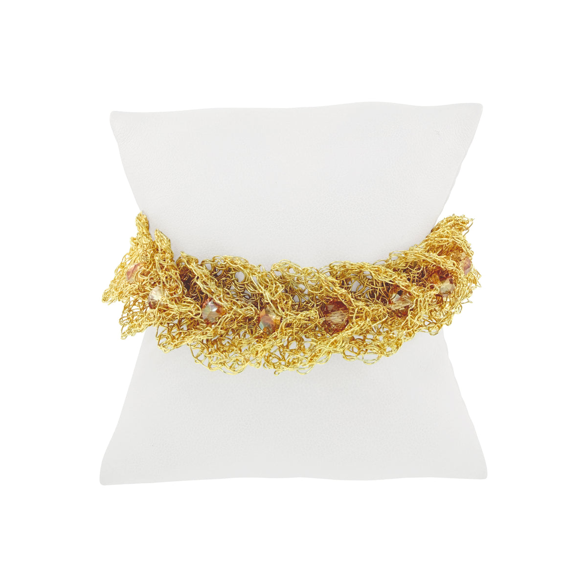 A gold-toned beaded and woven bracelet displayed on a white square jewelry stand against a white background. Ambar & Gold Calla Embrace handmade crochet bracelet featuring sculptural golden wire ruffles, sparkling amber crystals, and an adjustable gold-tone sliding chain closure.