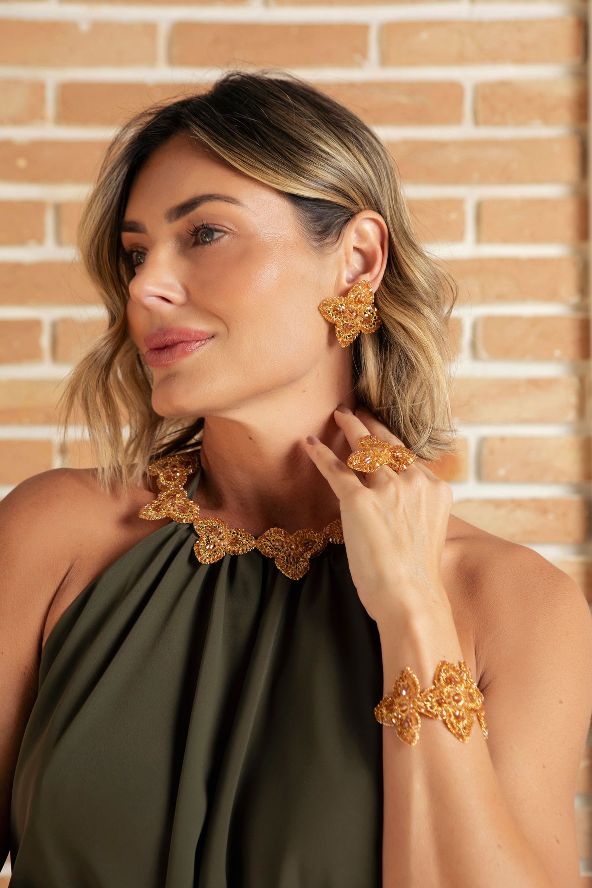 A woman wearing a matching gold jewelry set with a statement necklace, ring, and cuff bracelet, styled with an olive green halter top against a brick wall background. Shop the Ambar & Gold Orchid Crescent Necklace, handmade in Brazil with golden floral motifs, amber crystals, and 18K gold-filled wire. Elegant statement jewelry.