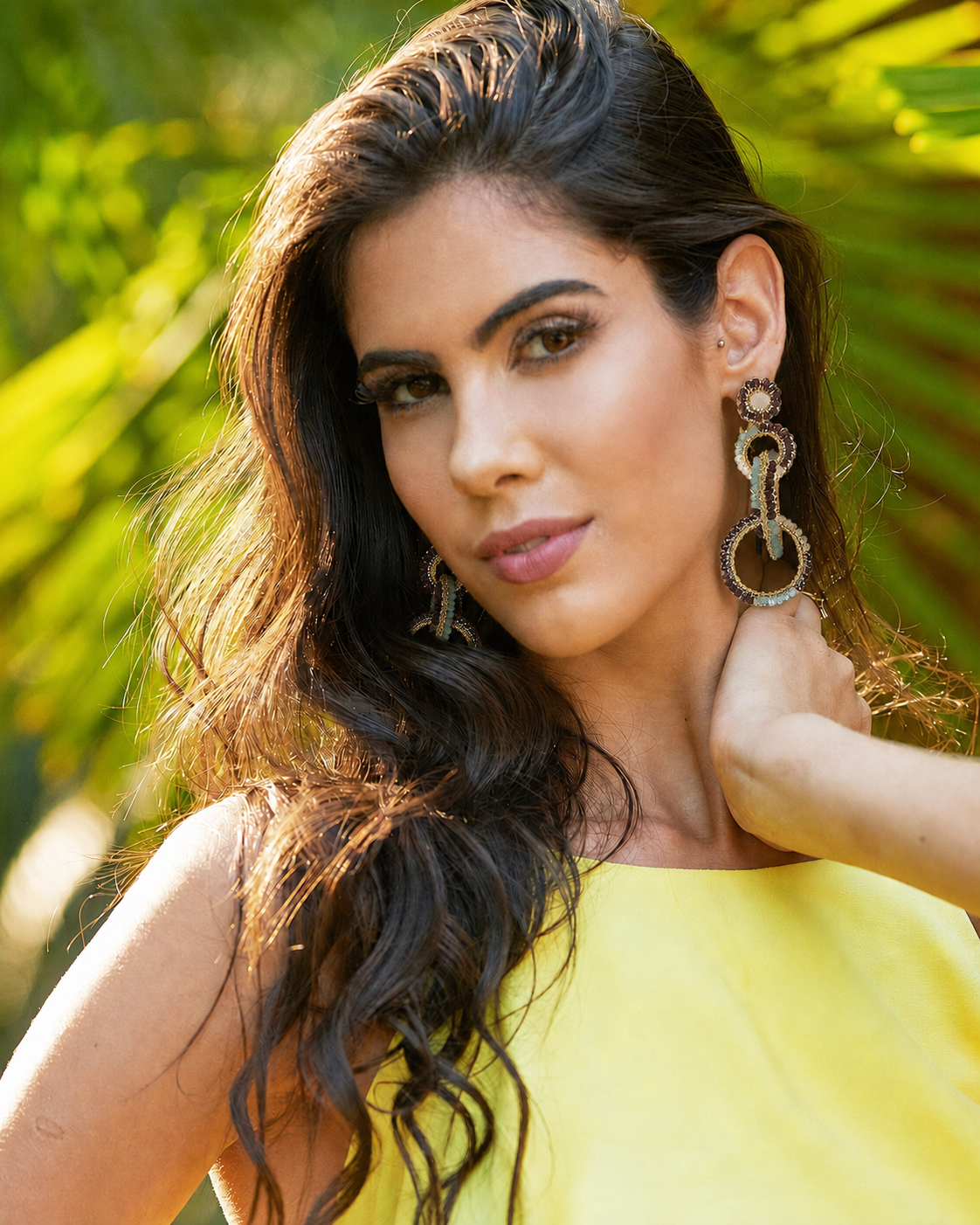 A woman with long, wavy dark brown hair wearing large, ornate silver-toned drop earrings and a bright yellow top, posing outdoors with a blurred background of green palm leaves. Shop interlocked link earrings handmade in Brazil with 18 karat gold filled wire, soft blue accents, ivory tones, and rich chocolate-brown beads. Lightweight, hypoallergenic, nickel-free, and artisan crafted.
