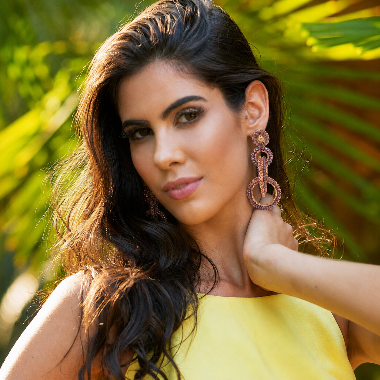 A woman with long dark wavy hair wearing a bright yellow top and large rose gold geometric drop earrings, posing outdoors with a blurred green palm leaf background. Shop interlocked link earrings handmade in Brazil with 18 karat rose gold filled wire, blush, mauve, and lavender bead accents, and sparkling crystals. Lightweight, hypoallergenic, and nickel-free.