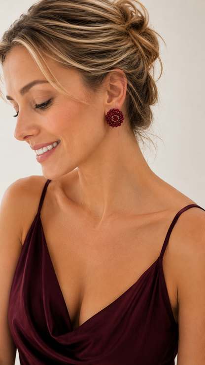 A woman with blonde hair in an updo wearing a dark burgundy spaghetti-strap top and a matching dark burgundy flower-shaped stud earring, shown in a close-up profile against a light neutral background. Burgundy & Gold Ava handmade crochet post earrings with faceted burgundy crystals and 18K gold filled wire in a floral design.
