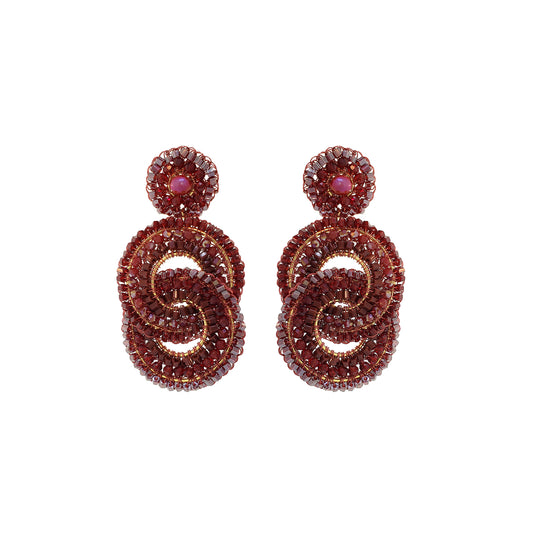 A pair of ornate drop earrings featuring a round stud with a pinkish-red center, connected to a larger double-loop spiral design paved with deep red faceted stones and accented by gold-toned metal. Discover Burgundy Loki Handmade Earrings, hand-crocheted in Brazil with 18K gold filled wire, sparkling crystals and glass beads in rich burgundy tones.