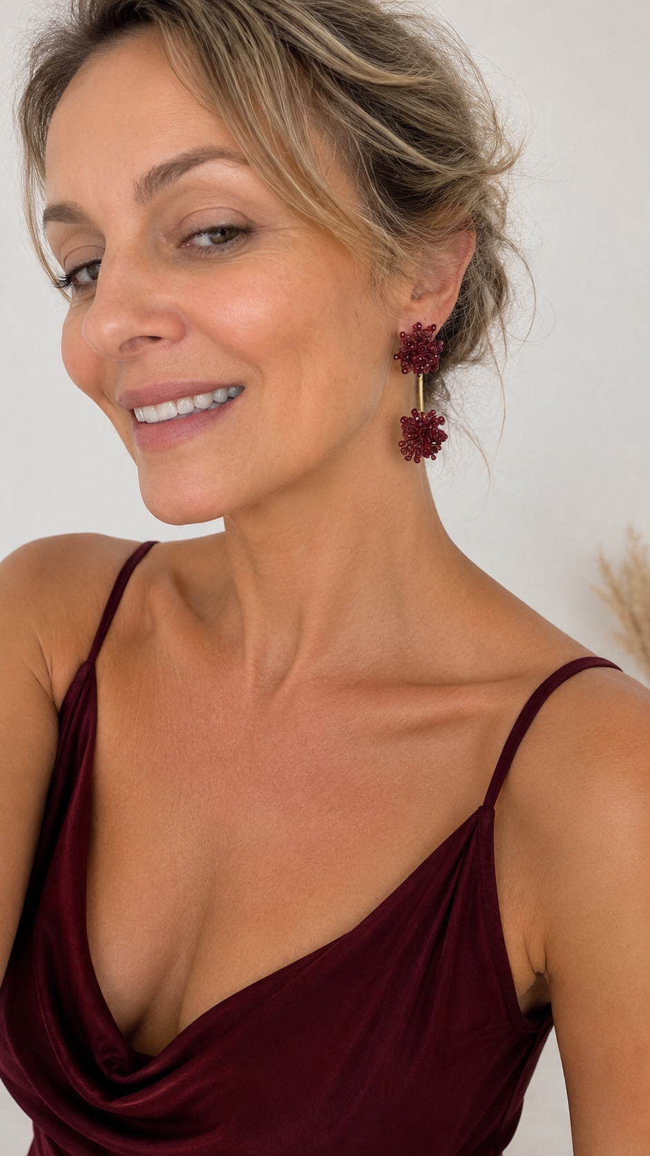 A close-up of a woman wearing a pair of dark red, textured flower-shaped drop earrings with gold-toned metal connectors, paired with a burgundy camisole top. Burgundy Mix Agatha Sprinkle handmade crochet earrings with two textured bead clusters connected by a sleek gold bar.