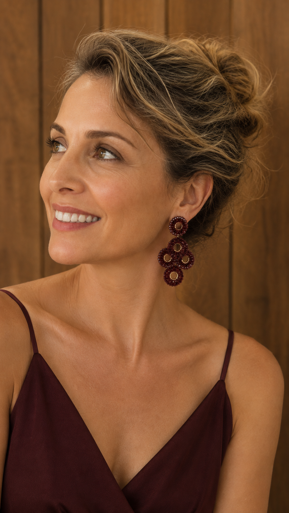 A woman with styled blonde hair in an updo wearing a dark burgundy spaghetti-strap top and a pair of large, dark red beaded drop earrings, set against a warm wood-paneled background. Burgundy Mix Coco handmade crochet chandelier earrings with layered burgundy crystal mandalas and 18 karat gold filled and enameled wire.