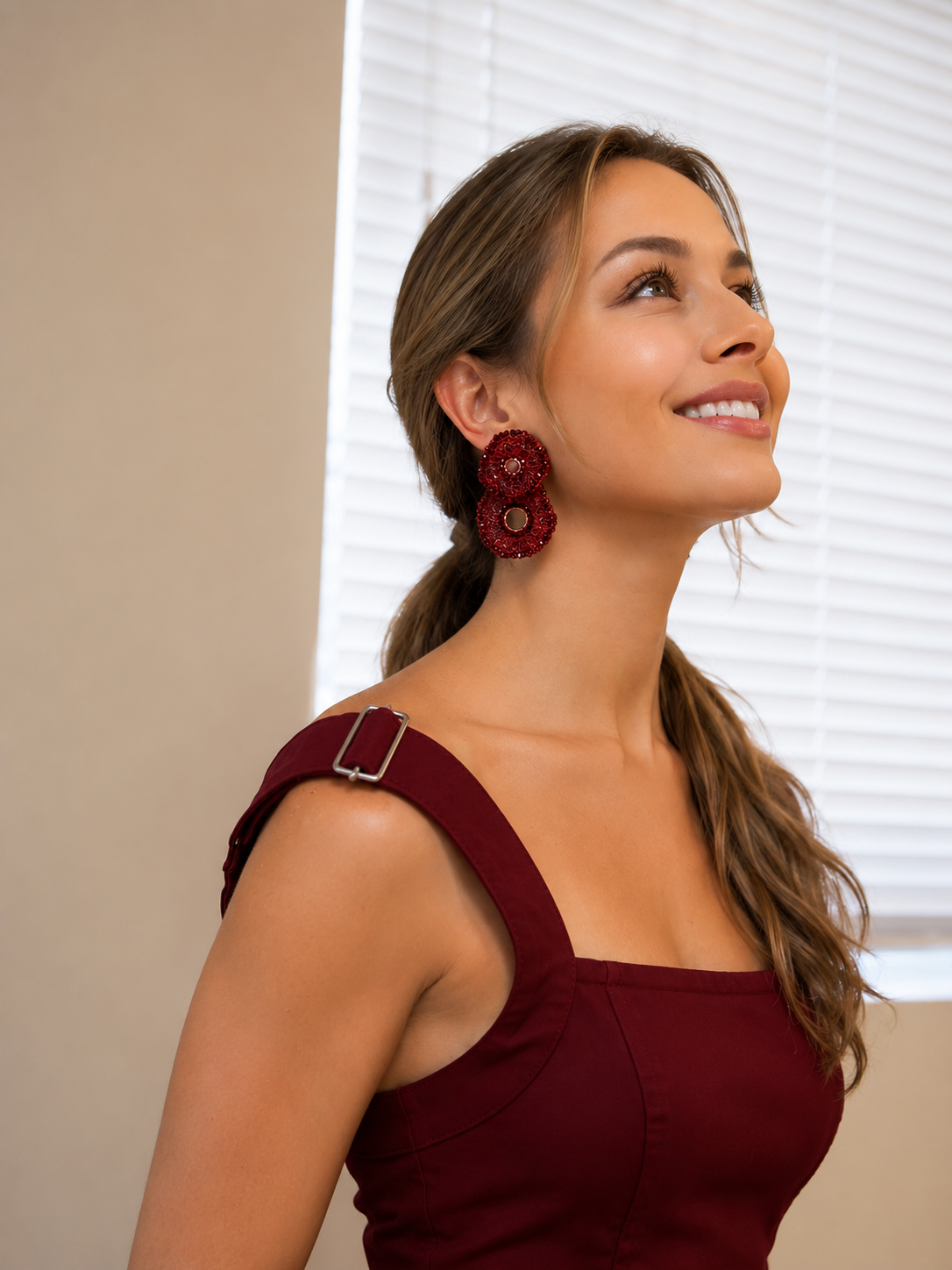 A woman with brown hair in a ponytail wearing a burgundy dress and a large, textured dark red flower-shaped earring, looking upward and smiling in front of white window blinds. Burgundy Mix Crista Duo handmade crochet earrings with layered circular shapes and shimmering bead accents, handcrafted in Brazil.