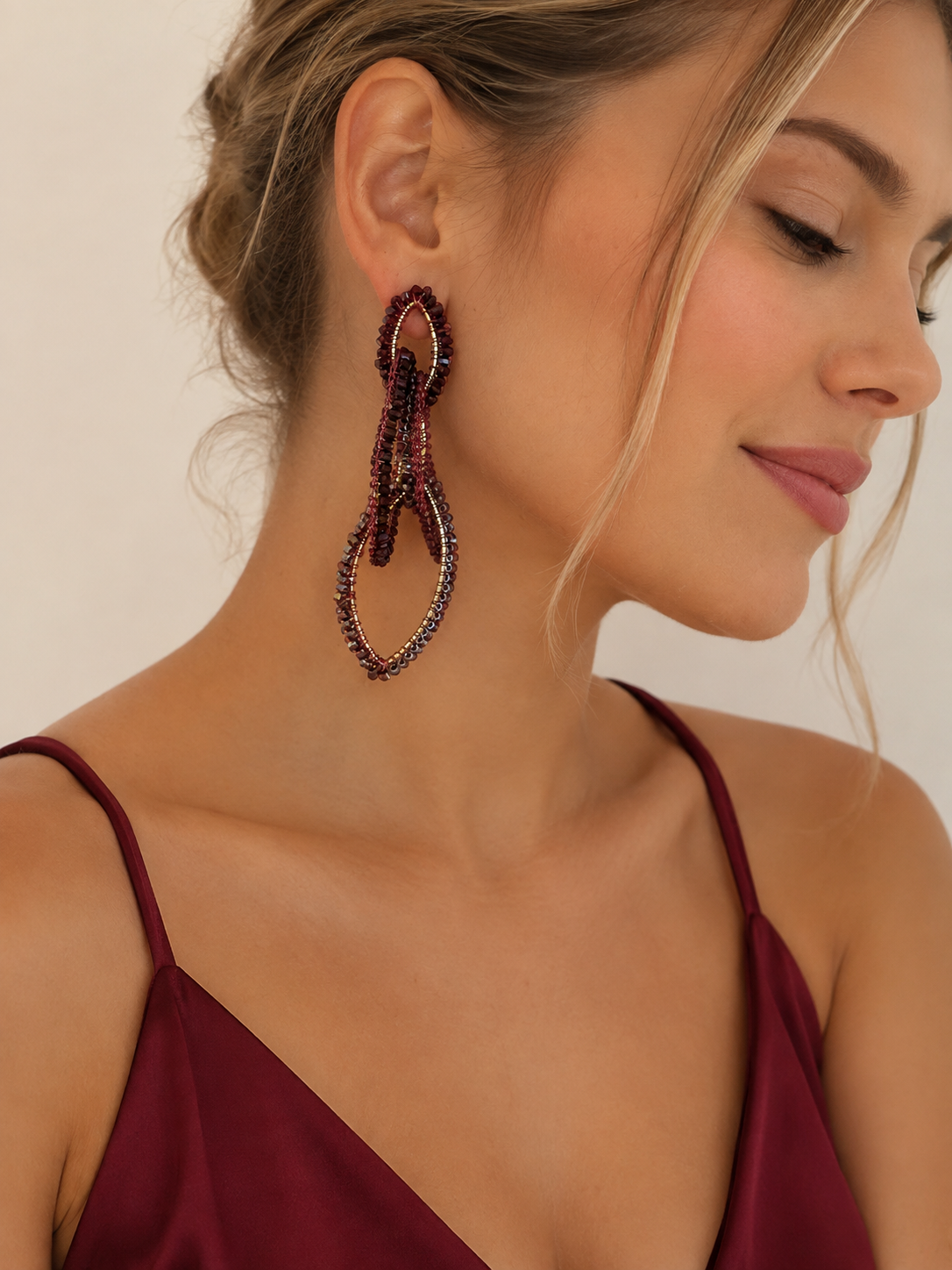 A close-up of a woman wearing a pair of large, dangling beaded earrings in deep burgundy and dark purple tones, paired with a matching burgundy camisole against a warm beige background. Burgundy Mix Demi handmade crochet dangle earrings with three cascading oval links, bead accents and intricate enameled copper wire.