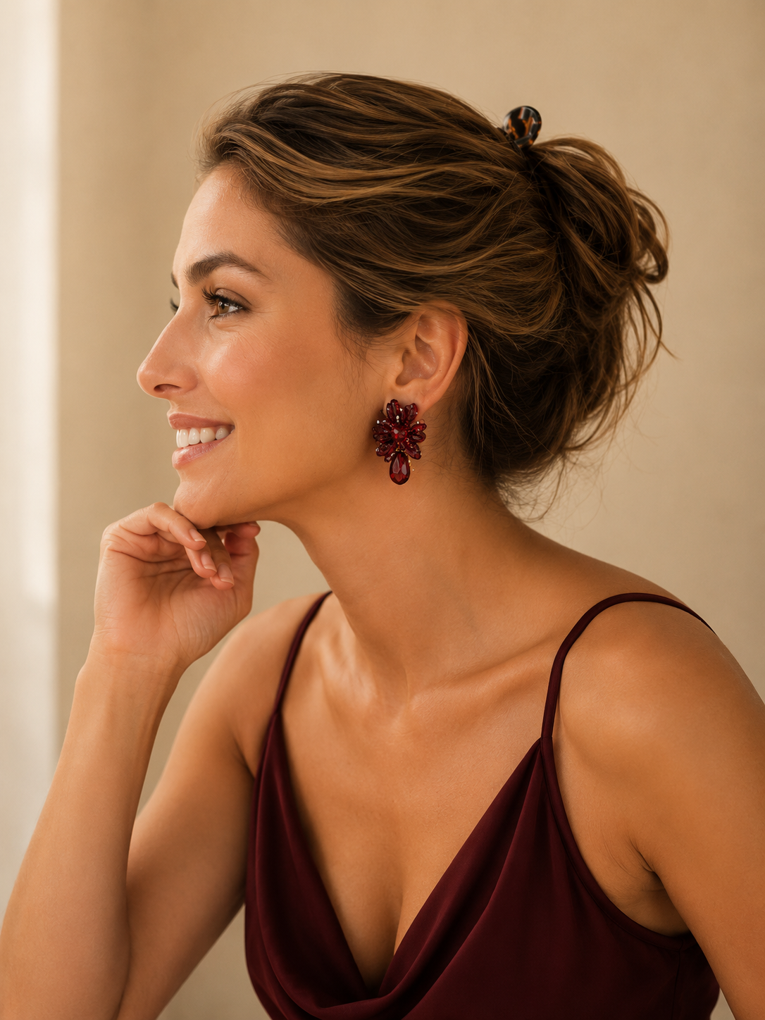 A woman in profile wearing a dark burgundy spaghetti-strap top and a matching hair clip, featuring a prominent dark red floral drop earring. Burgundy Mix Drizzle Mini Post handmade crochet earrings with textured burgundy bead clusters and a faceted crystal teardrop.