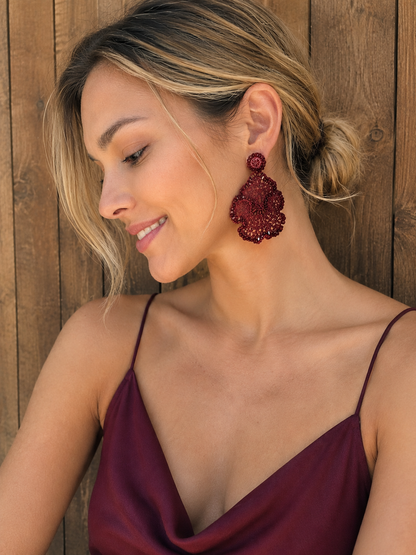 A woman with blonde-highlighted hair wearing a large, textured deep red beaded dangle earring, posing against a rustic wooden plank wall. Shop the Burgundy Mix Jasmine Trio Earrings, featuring sculptural hand-crocheted layers with shimmering bead accents. Lightweight and handmade in Brazil.