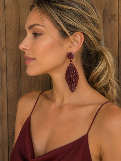 A woman in profile wearing a large, textured burgundy beaded dangle earring, set against a rustic wooden plank background. Shop Burgundy Mix Leaf Earrings, hand-crocheted in Brazil with shimmering beads and crystals in a sculptural leaf design. Lightweight and hypoallergenic.