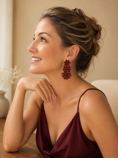 A woman with her hair in an updo is shown in profile, wearing a dangling cluster earring made of dark red beads. She is wearing a burgundy satin camisole top and resting her chin on her hand in a warm, softly lit indoor setting.