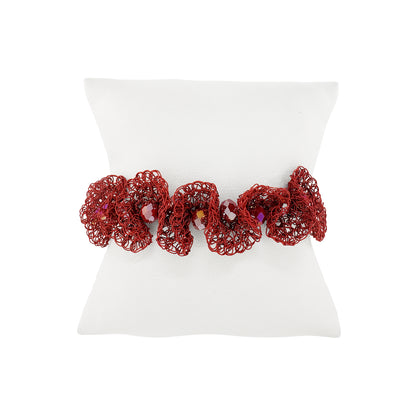 A red crochet hair scrunchie with small bead accents, displayed on a white pillow against a white background. Shop the Burgundy Mix Nalu Bracelet, handcrafted in Brazil with 18K gold-filled and enameled wire in a lightweight, adjustable statement design.