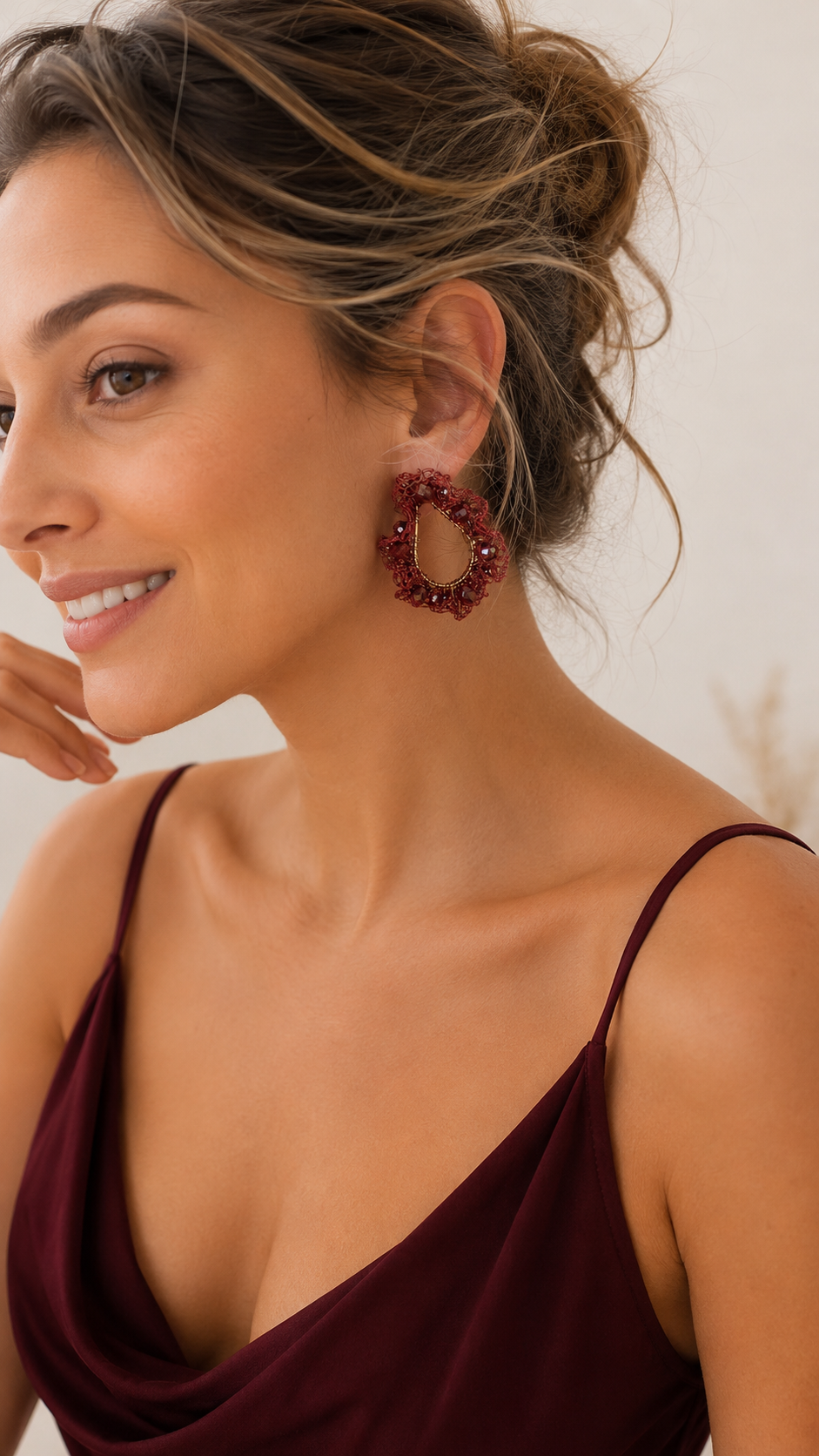 A close-up lifestyle shot of a woman wearing a pair of chunky, textured circular drop earrings in a deep burgundy red color. Burgundy Mix Nalu handmade crochet teardrop earrings with ruffled wire details and sparkling faceted burgundy bead accents.