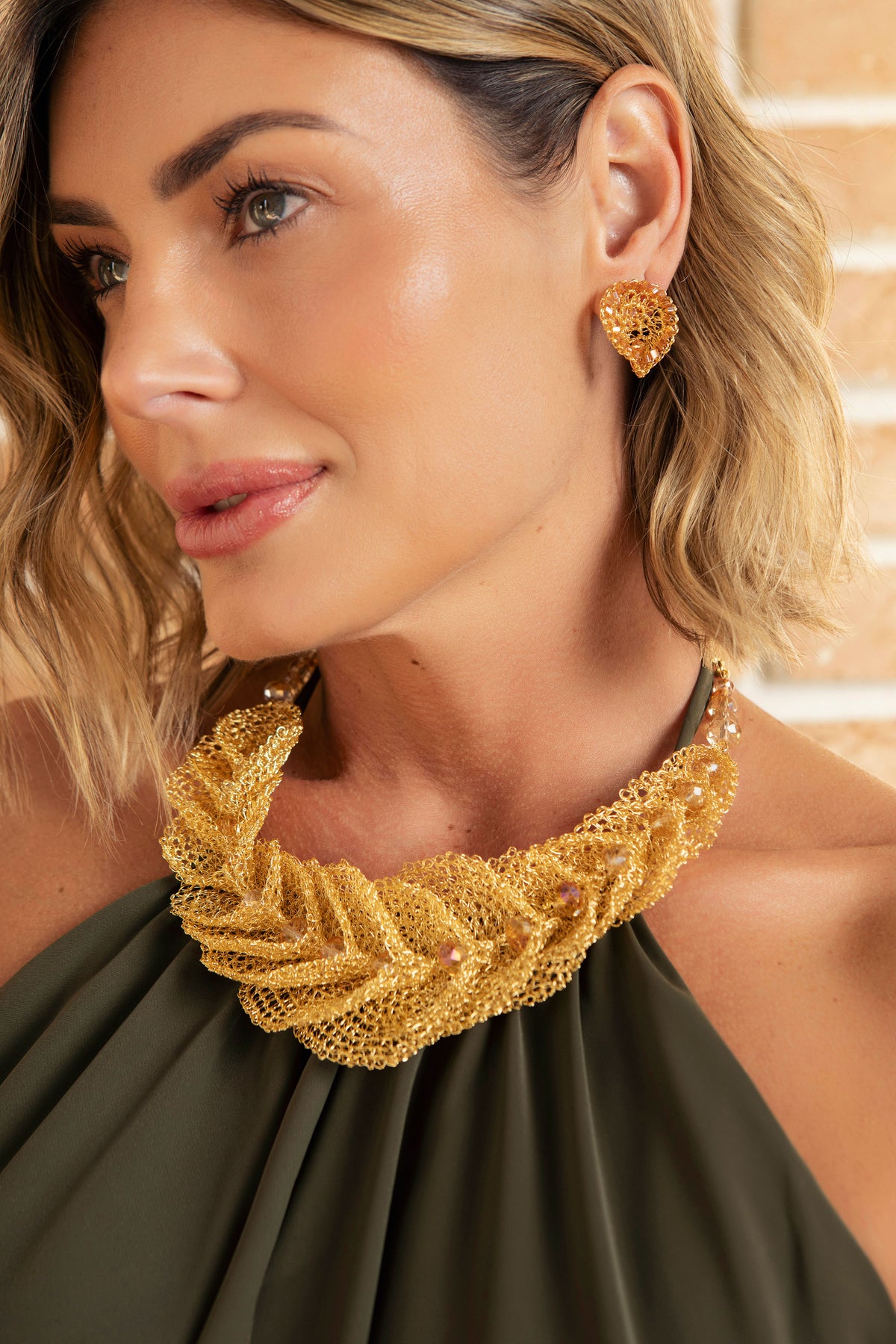 A close-up lifestyle shot of a woman wearing a chunky, textured gold statement necklace and matching gold heart-shaped stud earrings, paired with an olive green halter top. Discover the Cala Embrace Maxi Necklace, handcrafted in Brazil with 18K gold-filled wire, sculptural golden ruffles, and sparkling crystals. Luxury statement jewelry.