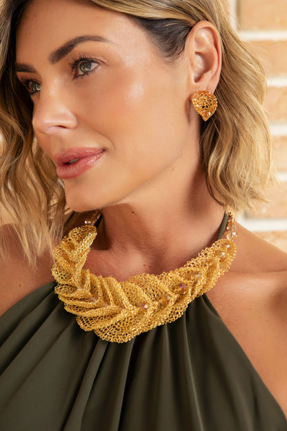 A close-up lifestyle shot of a woman wearing a chunky, textured gold statement necklace and matching gold heart-shaped stud earrings, paired with an olive green halter top. Discover the Cala Embrace Maxi Necklace, handcrafted in Brazil with 18K gold-filled wire, sculptural golden ruffles, and sparkling crystals. Luxury statement jewelry.