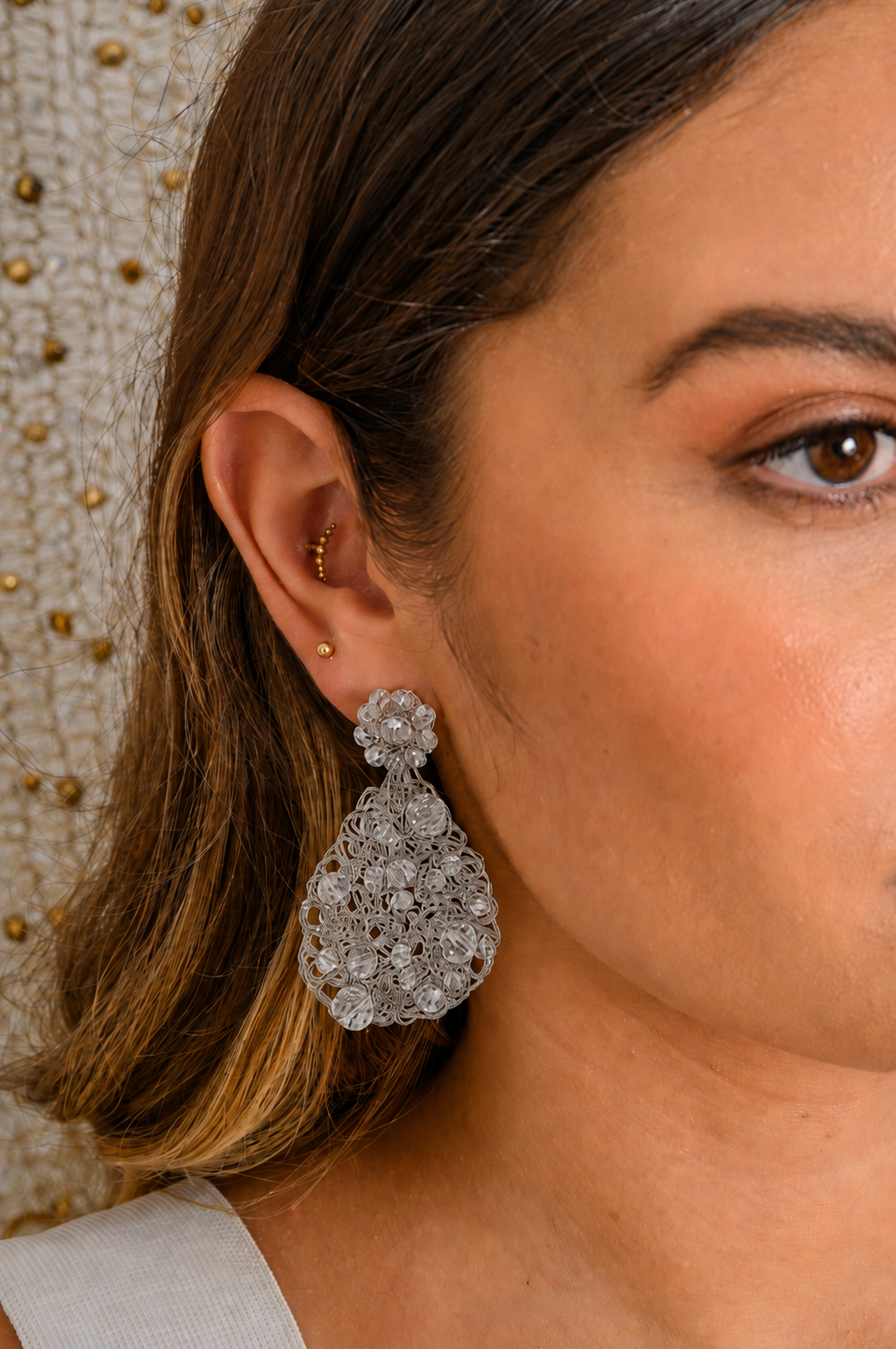 Close-up of a woman wearing a sparkling silver earring with a neutral background. Handmade in Brazil, these teardrop mesh earrings feature 18 karat gold filled wire, multicolor crystals, lightweight comfort, and hypoallergenic materials. 