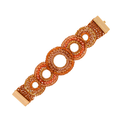 A beaded stretch bracelet with a warm orange, peach, and gold color palette, featuring a series of interlocking oval links with open centers and a light wood-toned clasp on a white background. Discover the Flamme Mix Gush Bracelet, handcrafted in Brazil with 18k gold-filled wire, vibrant orange crystals, and intricate crochet circle designs.