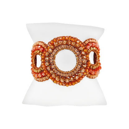 A wide beaded cuff bracelet displayed on a white mannequin stand. The bracelet features a large central oval surrounded by concentric rows of faceted beads in orange, peach, and gold, flanked by two smaller oval links on the sides. Flamme Mix Gush Bracelet featuring interlocking hand-crocheted circular motifs embellished with orange, amber, and champagne crystals, finished with a gold-tone magnetic clasp.