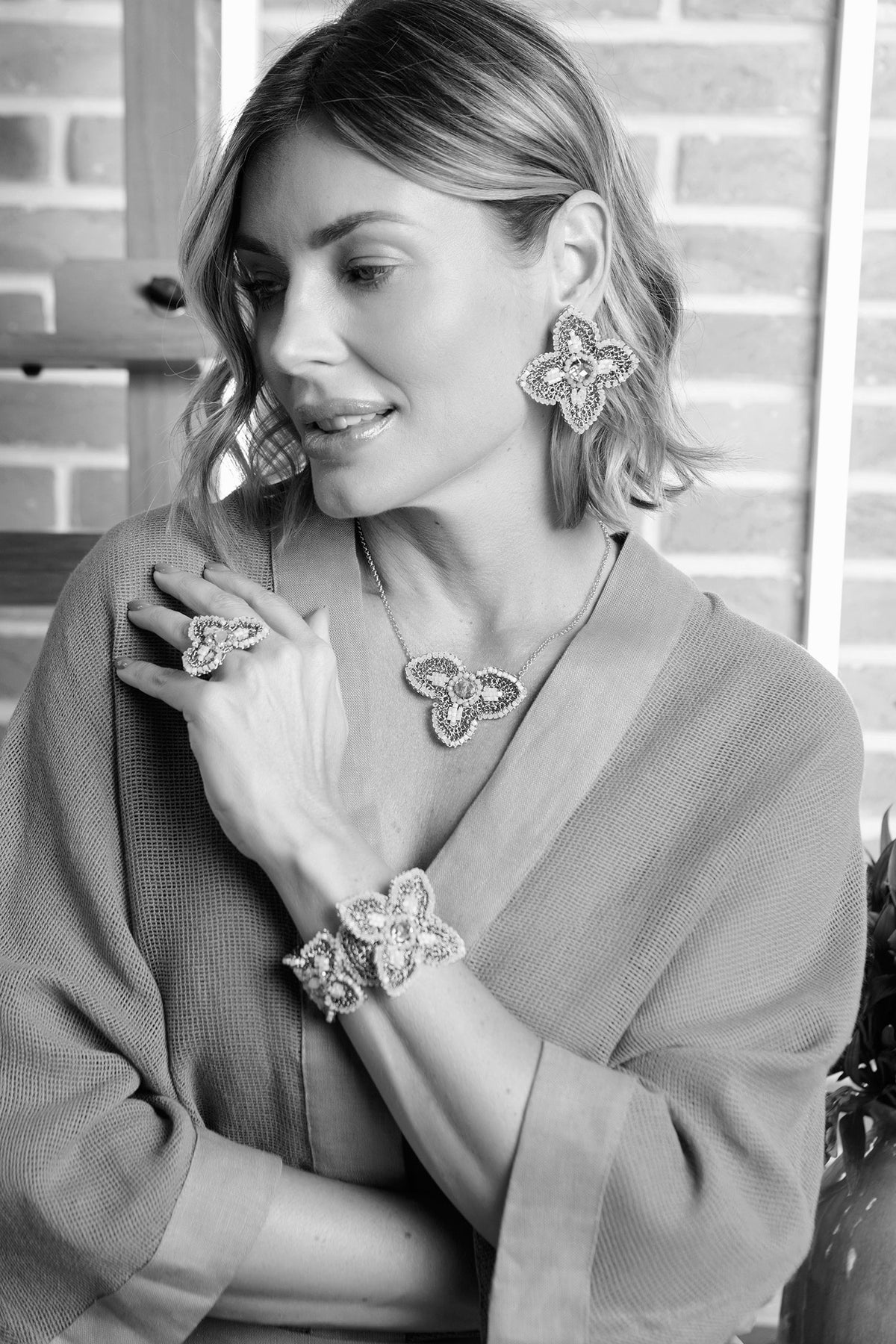 A woman wearing a matching jewelry set featuring a butterfly-shaped ring, necklace, and bracelet, along with large butterfly earrings. The image is in black and white, showing her in a textured cardigan against a brick wall background. Handmade orchid necklace, Flamme Mix necklace, floral pendant necklace, orange crystal necklace, Brazilian handmade jewelry, crochet pendant necklace, artisan statement necklace, Eclosion Collection, Lavish by Tricia Milaneze.