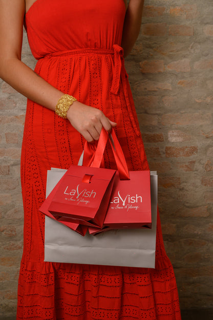Person wearing a red dress holding two Lavish shopping bags against a brick wall. Hand-crocheted 18k gold filled shell cuff bracelet handmade in Brazil, lightweight hypoallergenic nickel-free artisan jewelry with magnetic clasp.