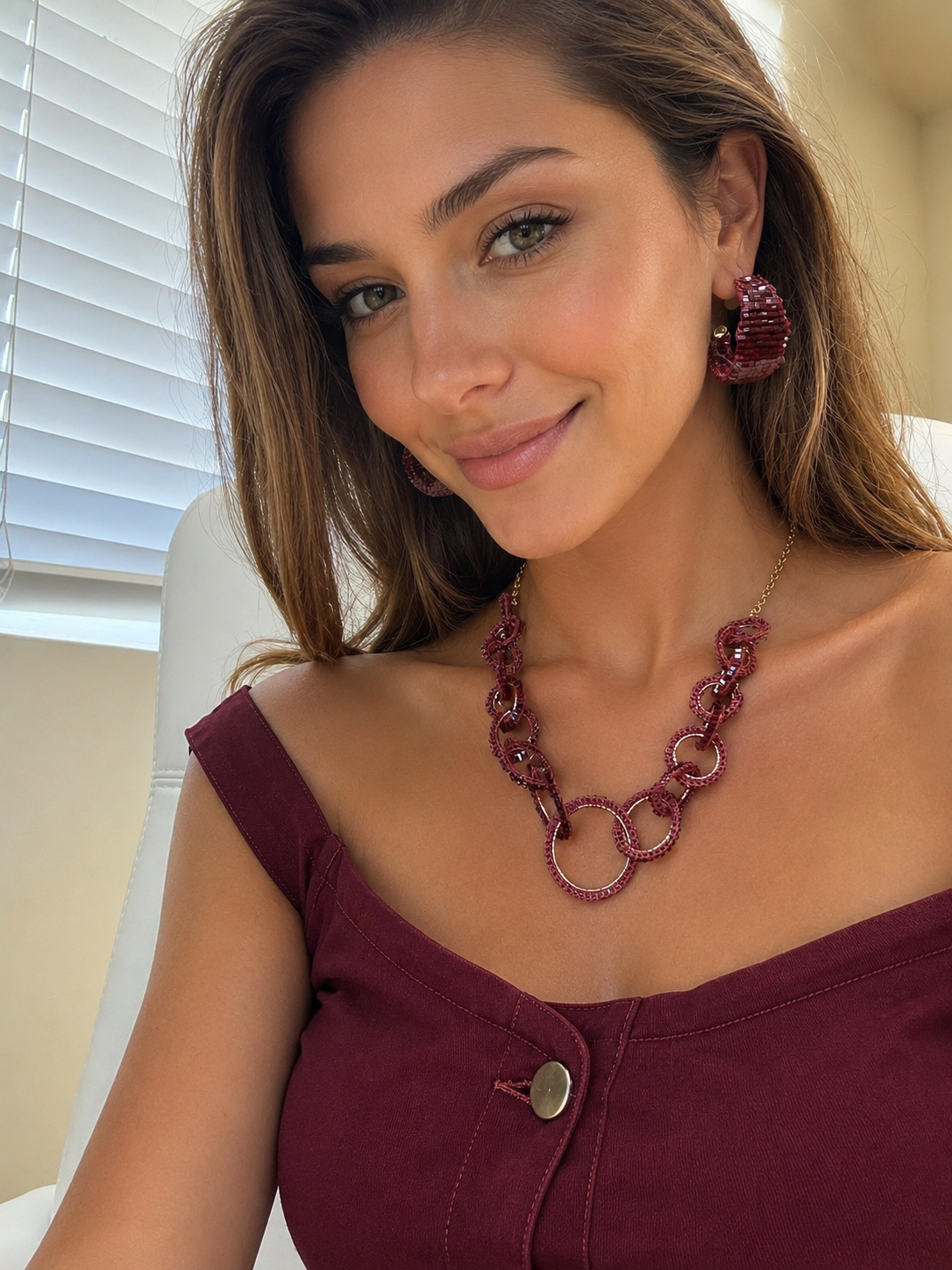 A woman wearing a chunky burgundy beaded chain link necklace with interlocking rings, styled with matching beaded earrings and a burgundy top. Lola handmade statement necklace with interlocking crochet links in burgundy enameled and 18K gold-filled copper wire.