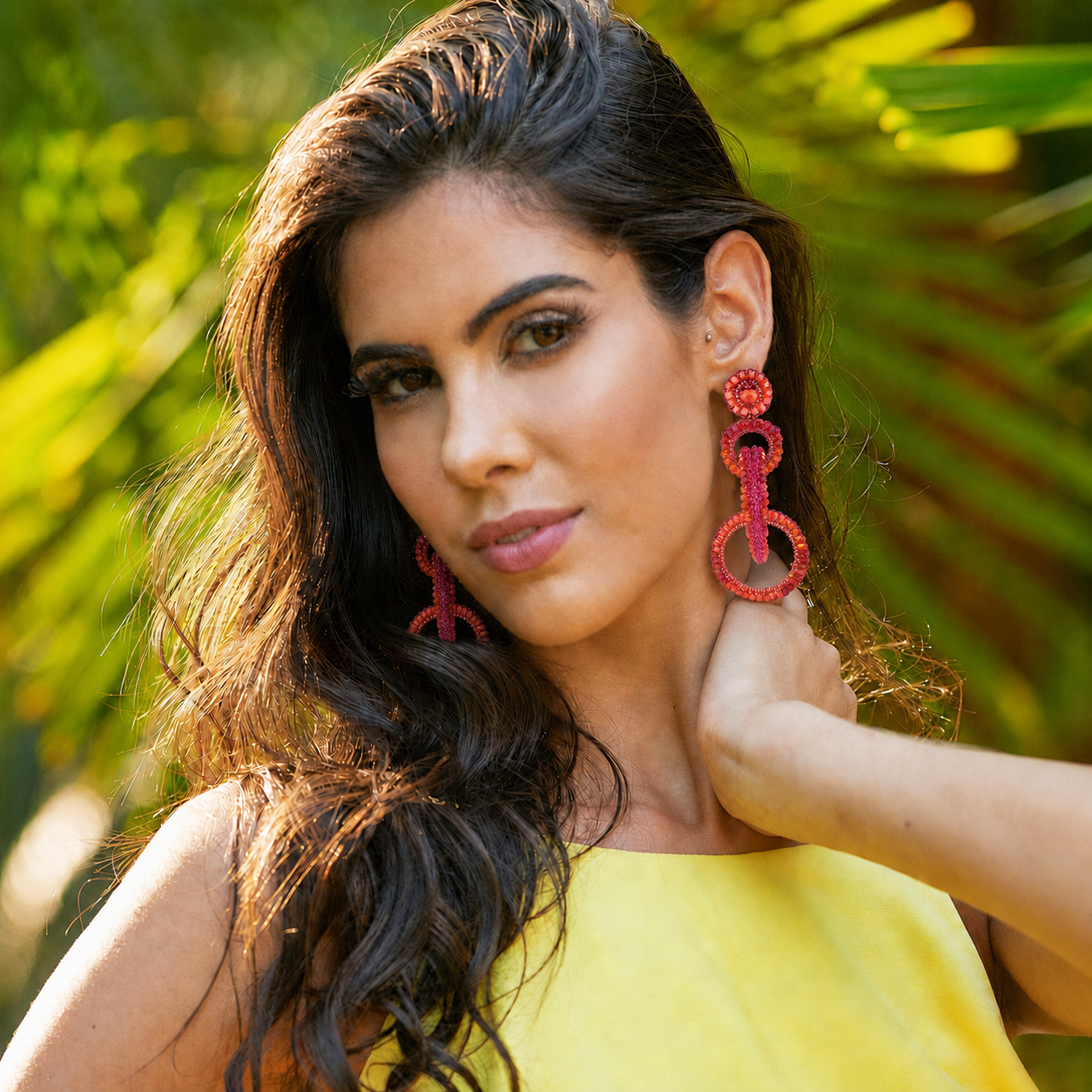 A woman with long dark wavy hair wearing large red beaded drop earrings and a yellow top, posing outdoors with a blurred green tropical background. Shop interlocked link earrings handmade in Brazil with bright fuchsia and orange enameled wire, sparkling bead accents, and layered circular links. Lightweight, hypoallergenic, nickel-free, and artisan crafted.