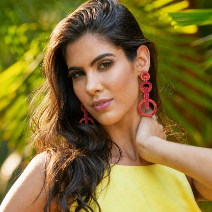 A woman with long dark wavy hair wearing large red beaded drop earrings and a yellow top, posing outdoors with a blurred green tropical background. Shop interlocked link earrings handmade in Brazil with bright fuchsia and orange enameled wire, sparkling bead accents, and layered circular links. Lightweight, hypoallergenic, nickel-free, and artisan crafted.