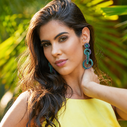 A woman with long dark wavy hair wearing large turquoise and purple beaded drop earrings, photographed outdoors with blurred green foliage in the background. Shop interlocked link earrings handmade in Brazil with neon turquoise enameled wire, sparkling crystal accents, and soft lavender and teal touches. Lightweight, hypoallergenic, nickel-free, and artisan crafted.