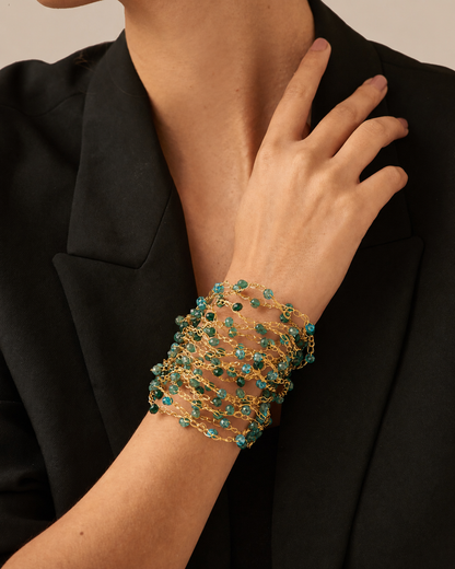 A close-up of a person wearing a wide, multi-strand bracelet with gold-toned metal and small teal-green faceted beads, styled with a black blazer. hand-crocheted cuff bracelet, gold cuff bracelet, green crystal bracelet, turquoise crystal bracelet, 18k gold filled bracelet, handmade Brazilian jewelry, artisan cuff bracelet, lightweight bracelet, hypoallergenic bracelet, nickel free bracelet, magnetic clasp bracelet, resort jewelry, vacation bracelet, jewel tone bracelet, statement cuff bracelet, luxury hand