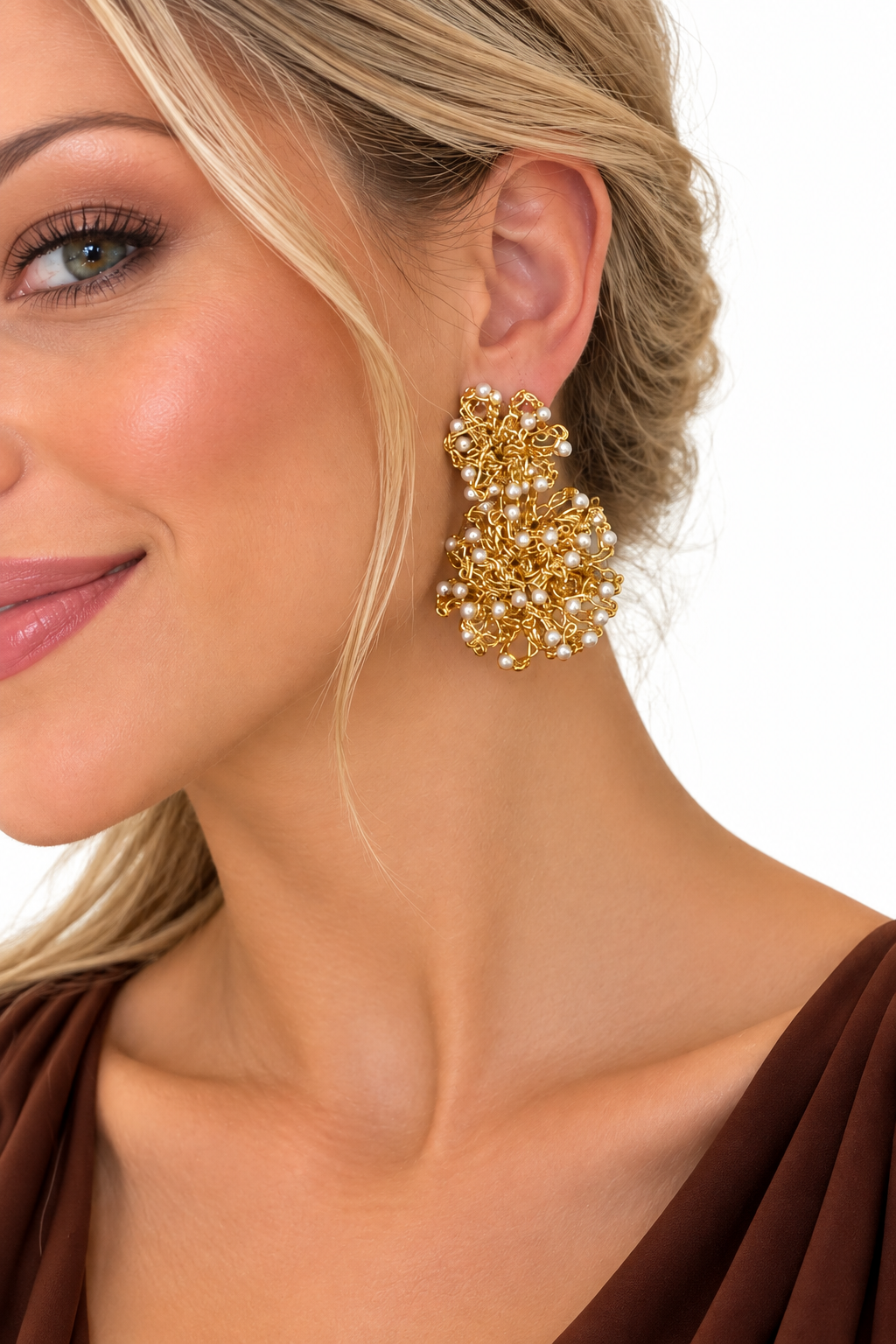 A close-up of a woman wearing a large, chunky gold statement earring adorned with clustered faux pearls, set against a clean white background. Shop Double Mandala Earrings, hand-crocheted in Brazil with 18K gold-filled wire and delicate pearl-like beads. Lightweight, hypoallergenic and artisan-made.