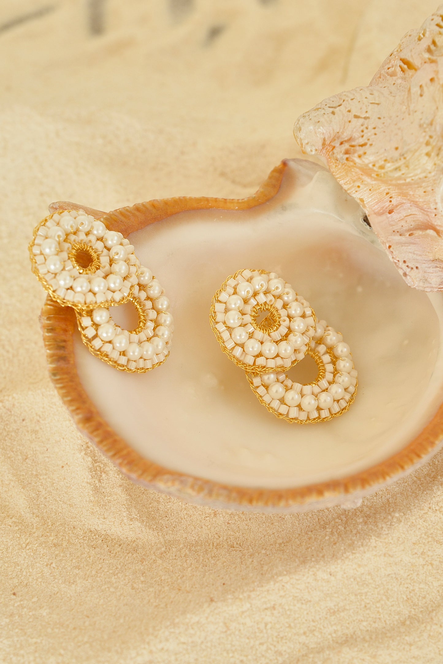 Pearl and gold earrings on a seashell with sand background