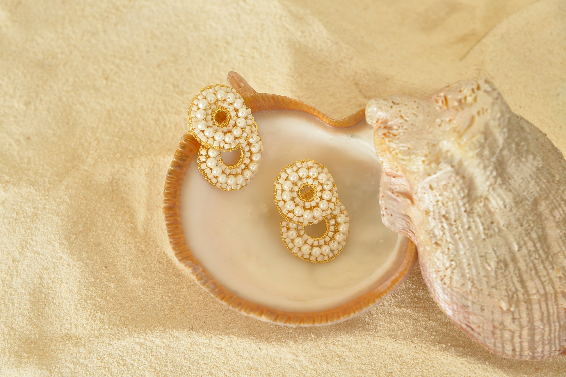 Pair of gold and pearl earrings on a shell with sand background