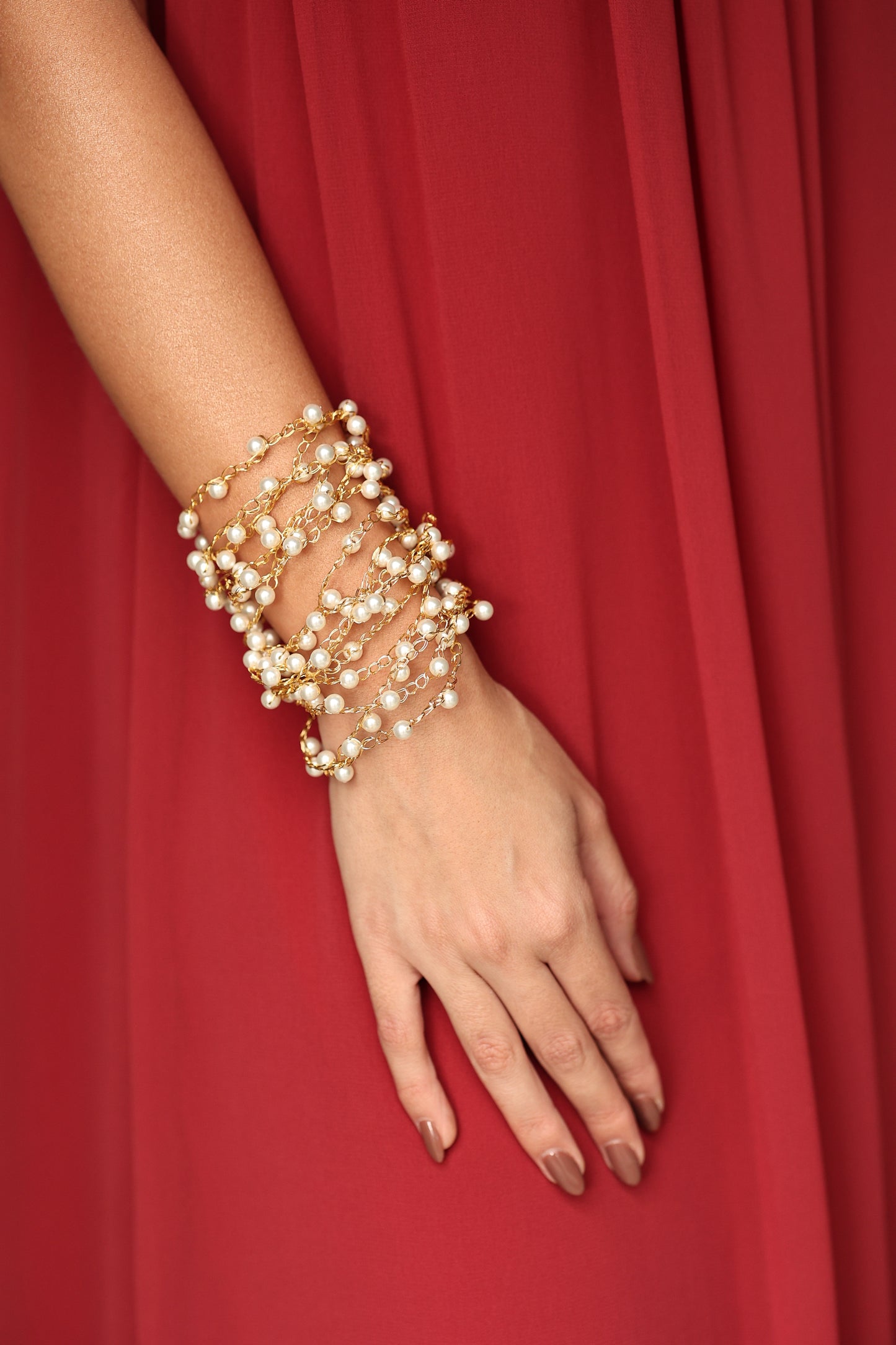 Hand wearing multiple pearl bracelets on a red fabric background. Shop a hand-crocheted cuff bracelet handmade in Brazil with 18 karat gold filled wire and delicate pearl accents. Lightweight, hypoallergenic, nickel-free, and finished with a magnetic clasp.