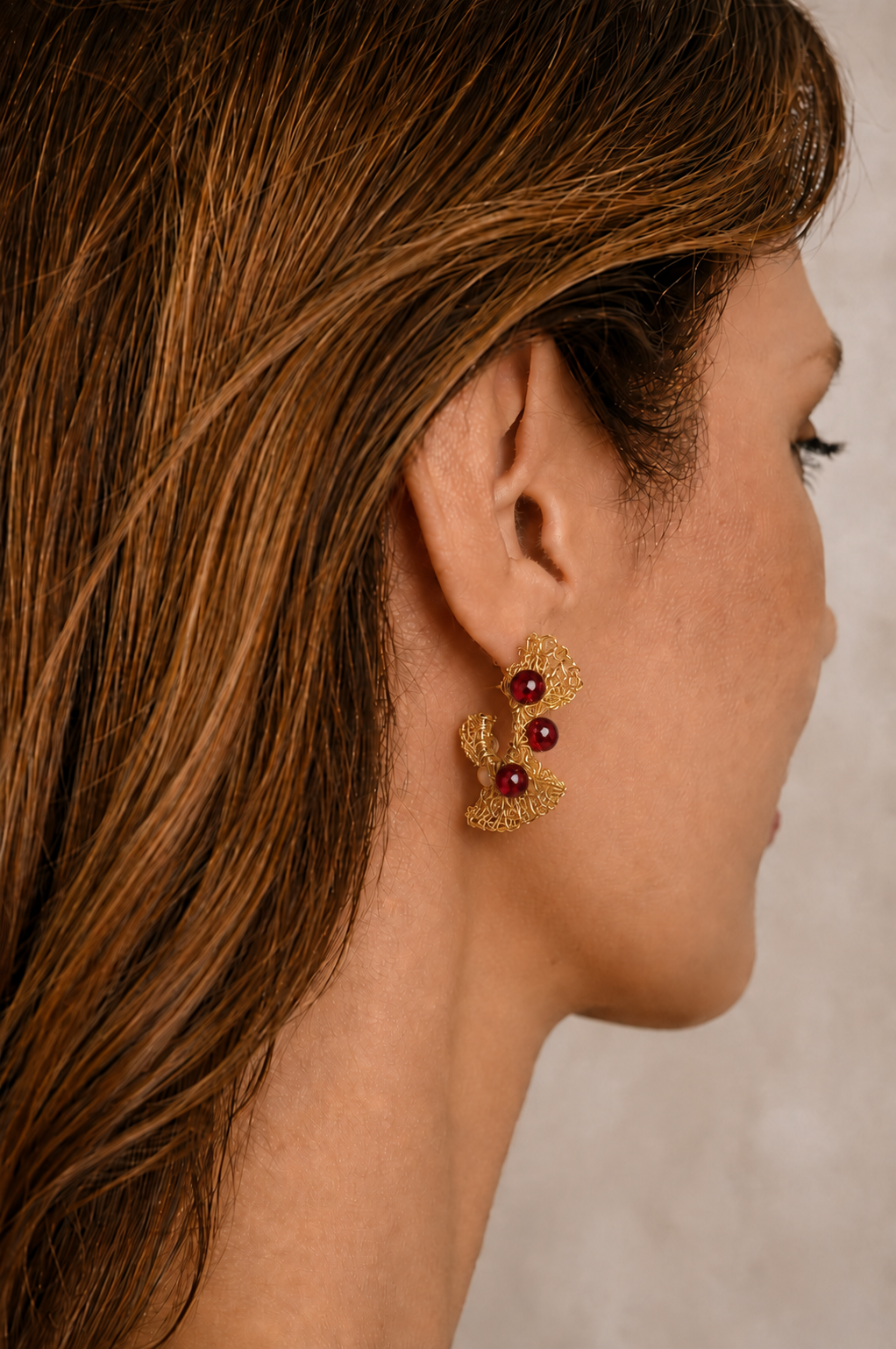 A close-up side profile of a woman with long brown hair wearing a gold-toned leaf-shaped earring with three small dark red pearl-like accents. Hand-crocheted 18k gold-filled hoop earrings with red crystals, handmade in Brazil by Lavish by Tricia Milaneze.