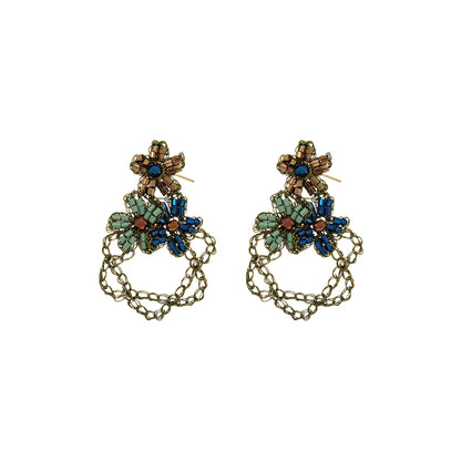 A pair of ornate dangling earrings with antique gold-tone chains and floral clusters of blue, green, and brown rhinestones, shown on a plain white background.  Shop Riviera Mix Aster Trio Earrings, handcrafted in Brazil with floral crochet details, crystals, beads and 18K gold-filled and enameled wire.