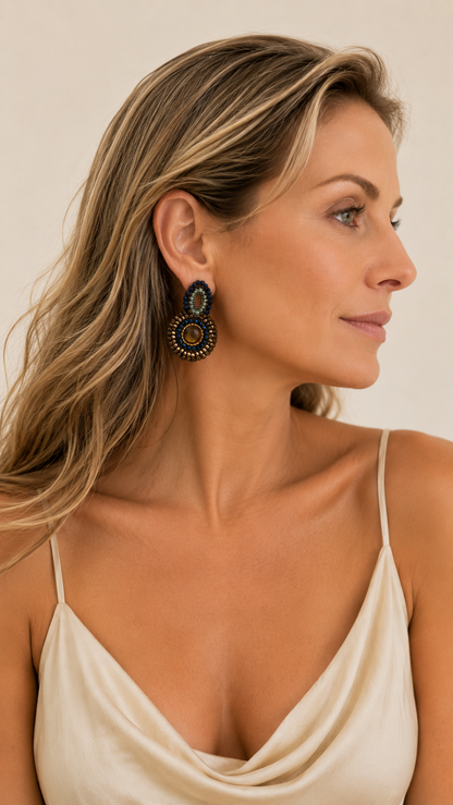 A woman in profile wearing a pair of large, dark circular statement earrings with gold and blue details, paired with a cream-colored spaghetti strap top against a warm beige background. Riviera Mix Billie Trio handmade crochet earrings with blue, sage green and bronze crystals, faceted center stones and 18 karat gold filled wire.