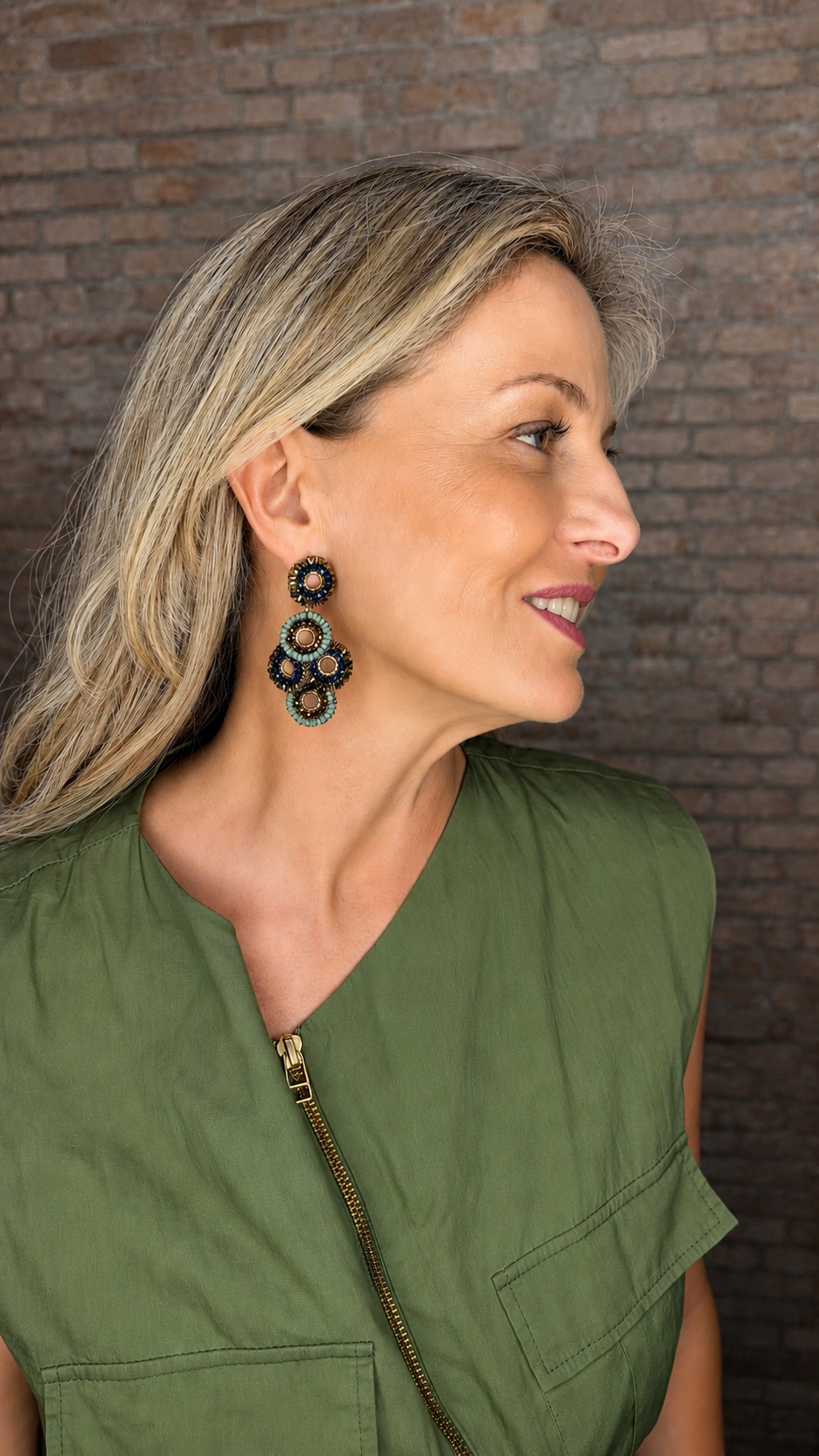 A woman in profile wearing a green sleeveless top and large, ornate black and gold statement earrings, set against a dark brick wall background. Riviera Mix Coco handmade crochet chandelier earrings with blue, sage green and bronze crystal mandalas and 18 karat gold filled wire.