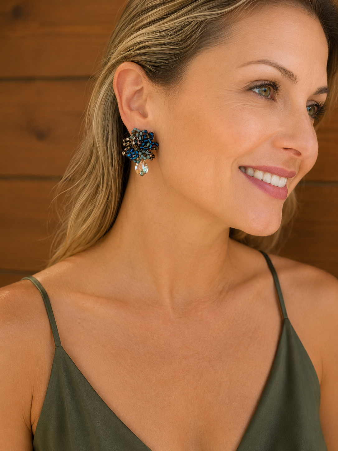 A close-up lifestyle shot of a woman wearing a pair of blue and silver beaded flower-shaped earrings, set against a warm wood-paneled background. Riviera Mix Drizzle Mini Post handmade earrings with blue, sage and bronze crochet bead clusters and a faceted crystal drop.