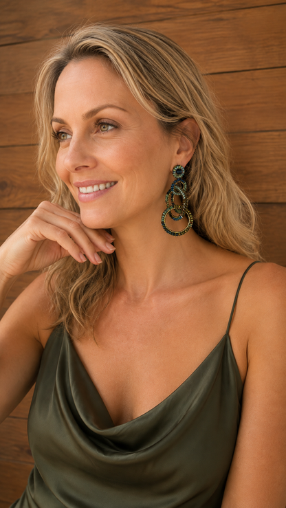 A smiling blonde woman wearing a dark olive green satin camisole top and large, multi-layered green and gold hoop earrings, posing against a rustic wooden wall. Riviera Mix Grace handmade crochet earrings with cascading interlocked rings in deep blue, sage green and bronze enameled wire.